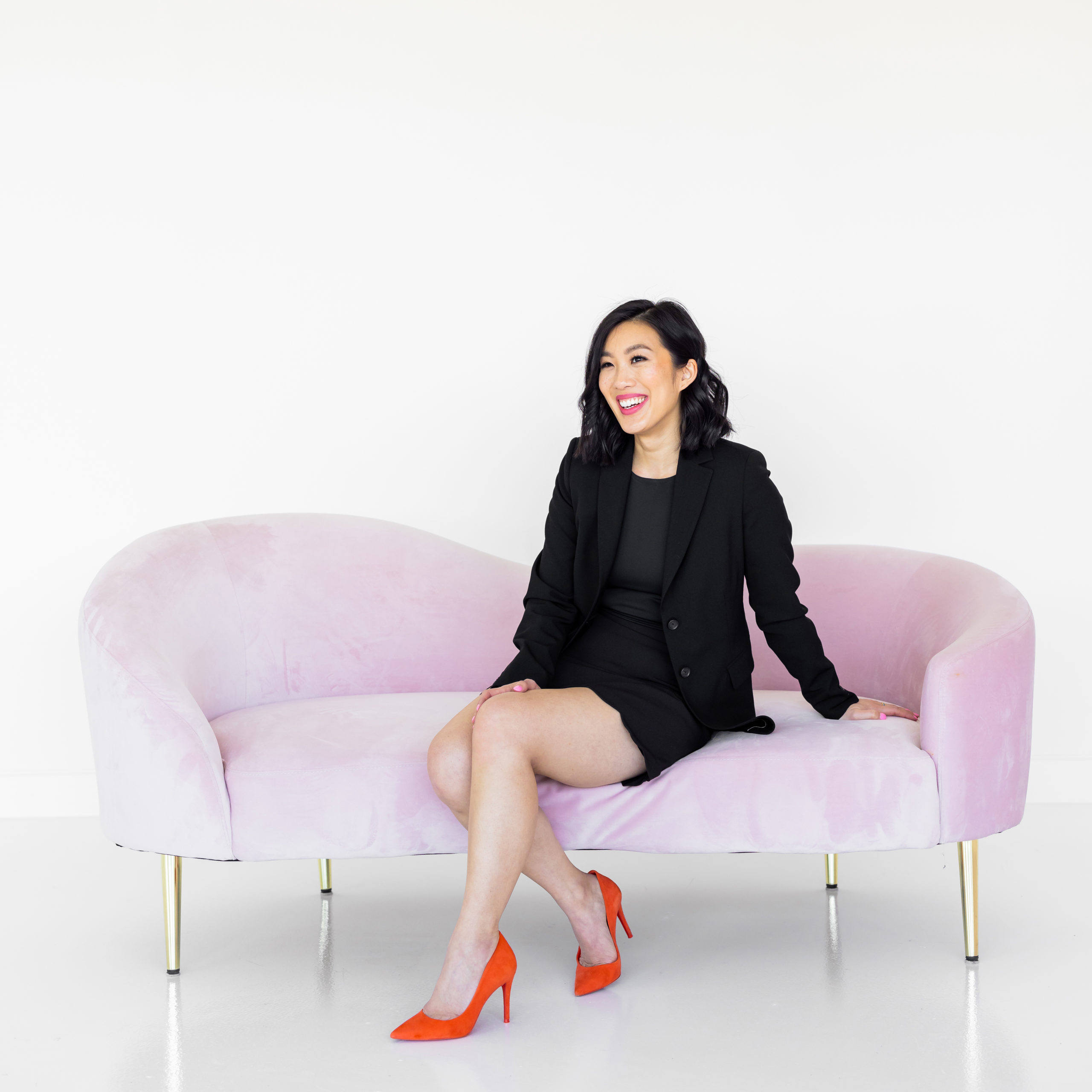 Asian woman in a black skirt and blazer and orange pumps sitting on a pink couch