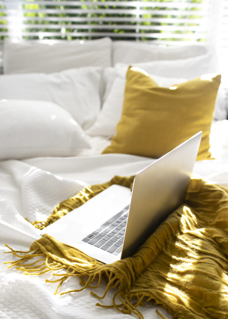 Open laptop on a sun-drenched bed with white sheets and yellow blanket