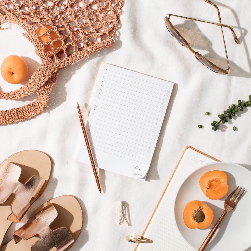 styled photo of a to do list notepad surrounded by gold sandals, plates of apricots, tortoiseshell sunglasses, and a woven tote bag