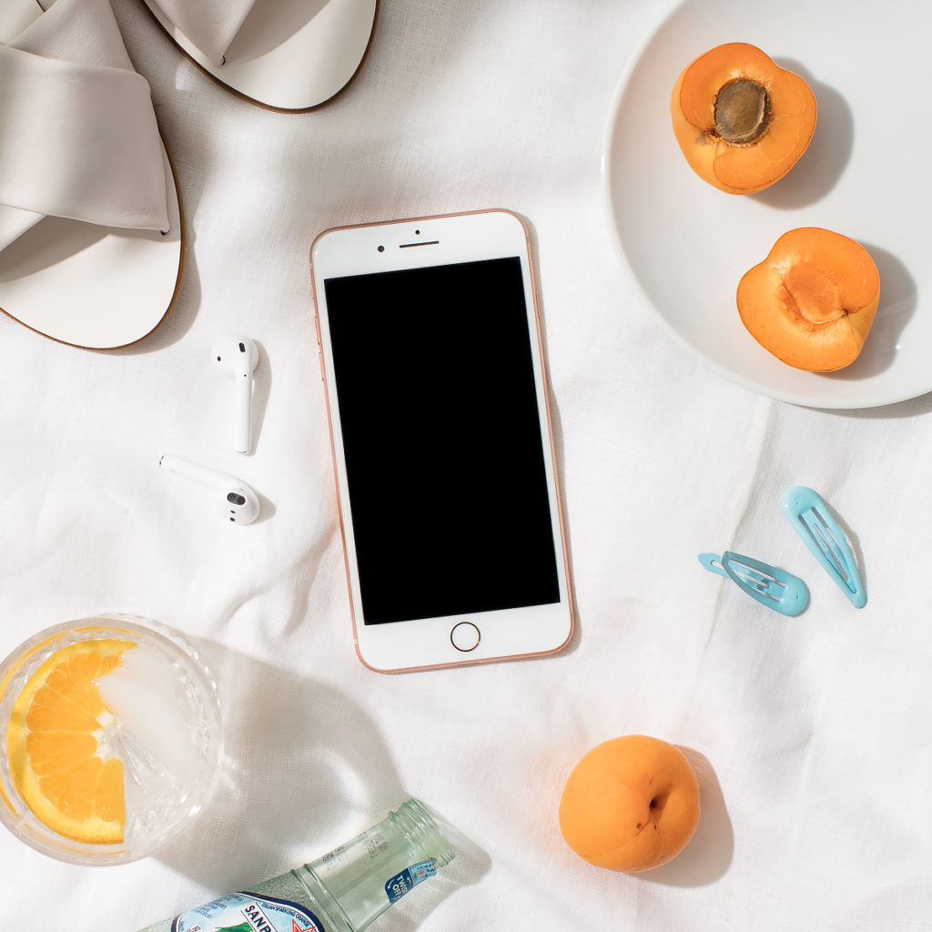 styled photo of a white iphone pro surrounded by white sandals, a plate of apricots, blue hair pins, and a glass of sparkling water