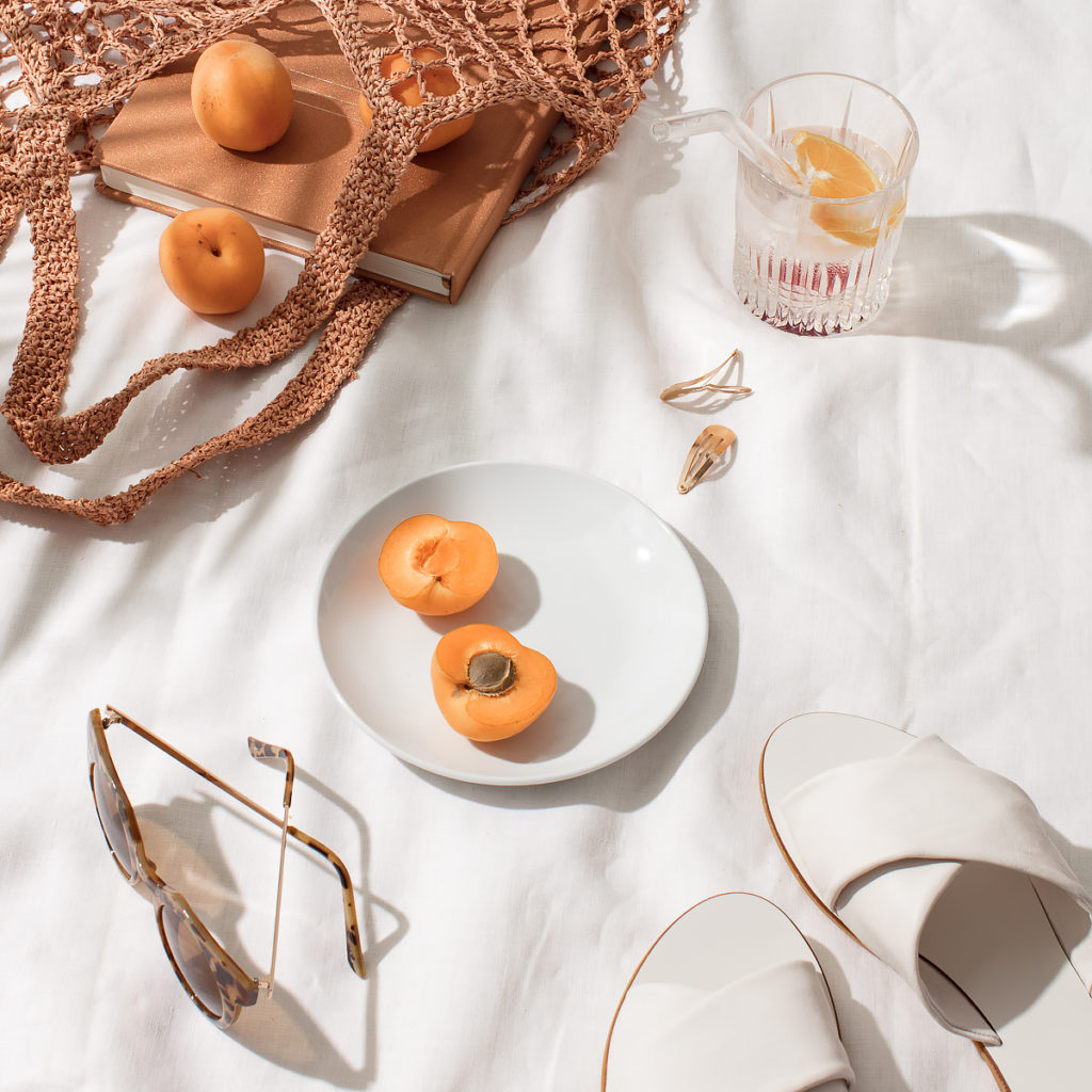 styled photo of a woven tote bag with tan books and apricots spilling from it, apricots on a plate, white sandals and tortoiseshell sunglasses, and a glass of water with a slice of orange.