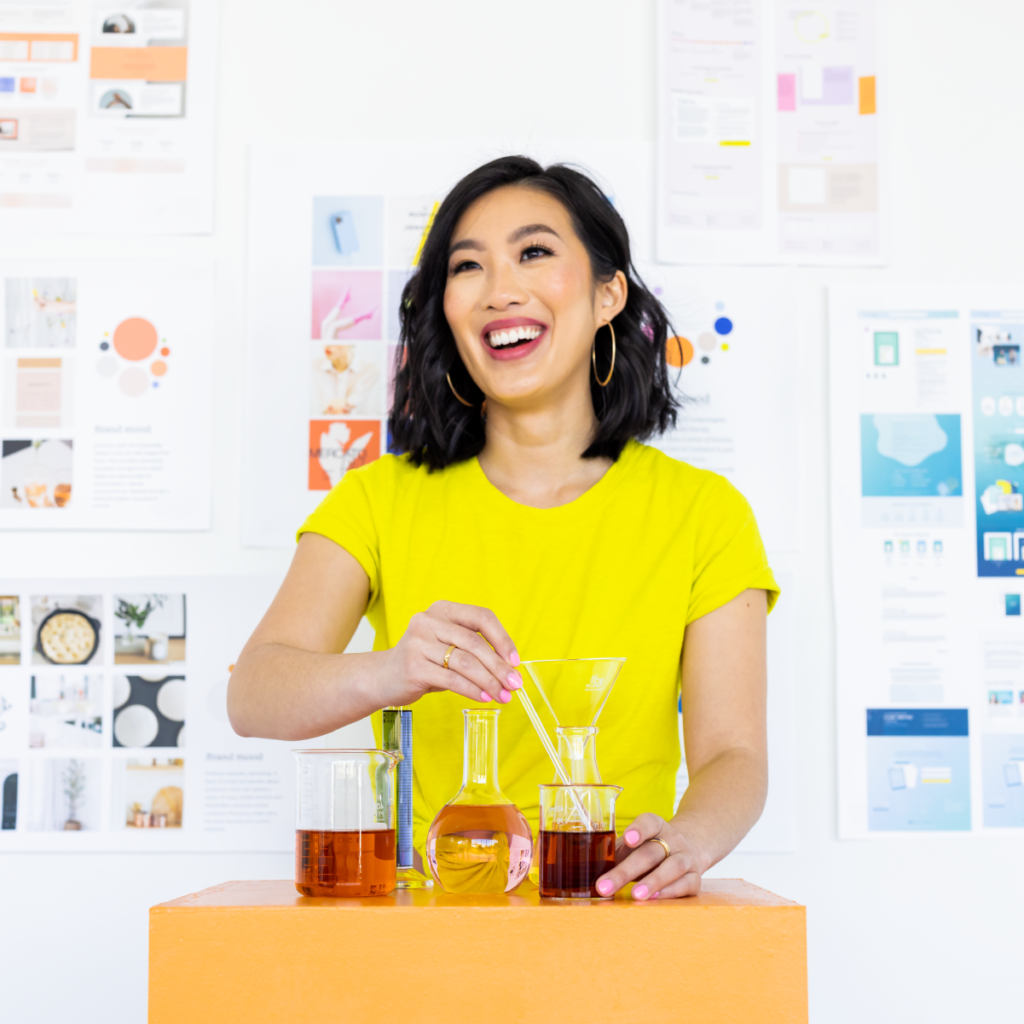 Asian woman wearing a yellow tshirt standing in front of a wall of brand mood boards, mixing beakers of colorful liquids.