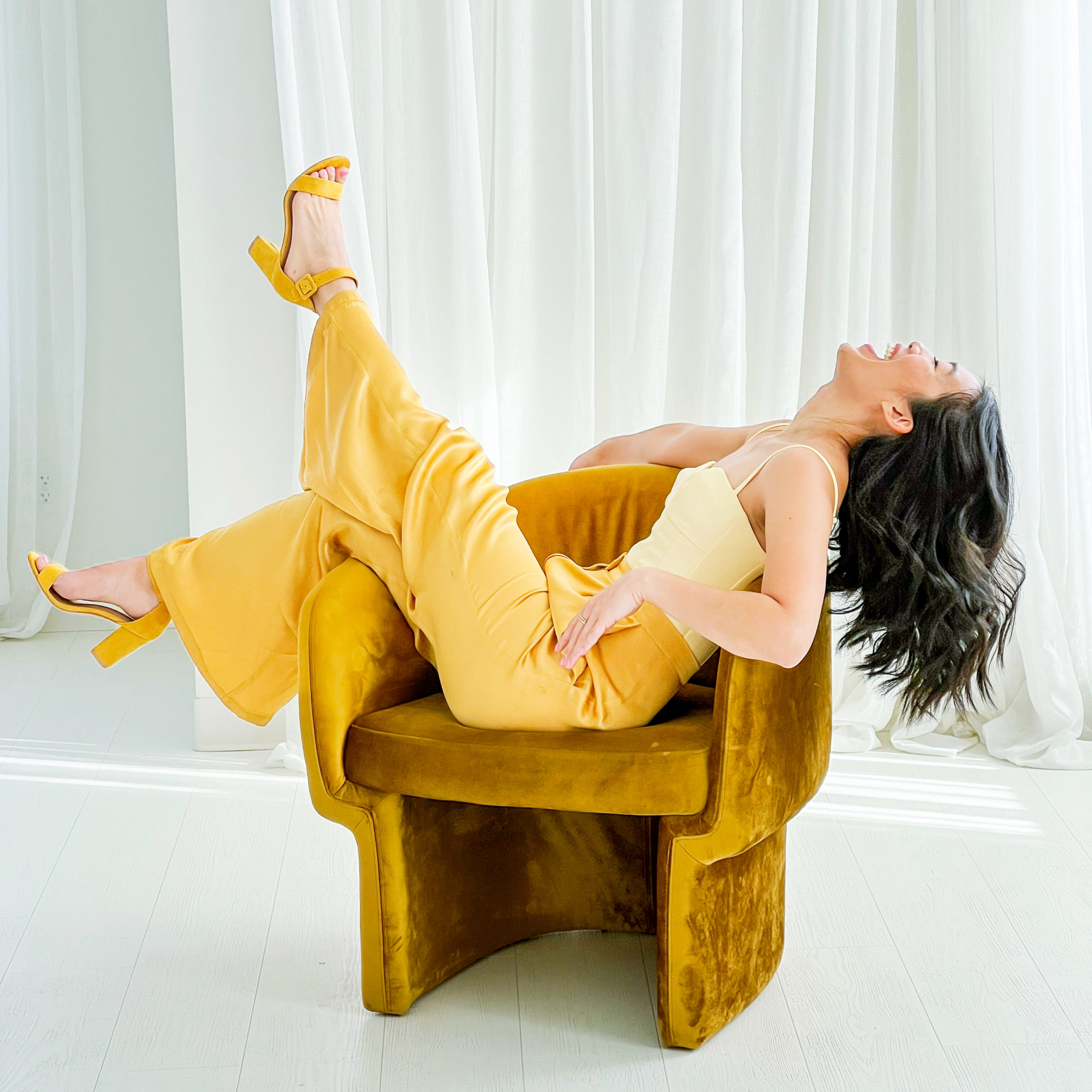 Image of Nicole Yang in a yellow bodice and yellow satin pants kicking her legs up in a yellow velvet chair